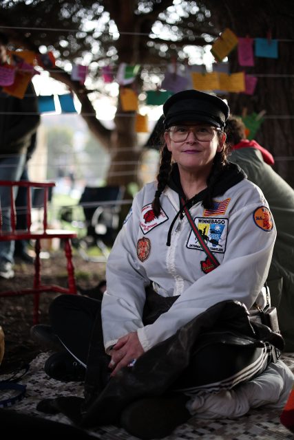 A person wearing a decorated white jacket and cap is seated outdoors, surrounded by colorful hanging notes and chairs in the background.
