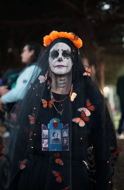 Person dressed in Day of the Dead costume with painted face, wearing a black veil adorned with butterflies and orange flowers, and a necklace with photos.