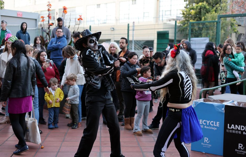 Dia de los Muertos celebration with two people in skeleton costumes dancing, surrounded by a crowd of onlookers.