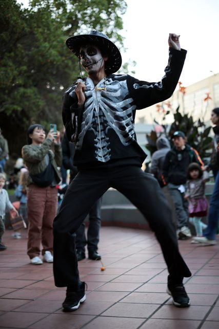 Person in skeleton costume and hat dancing in outdoor area, surrounded by people.
