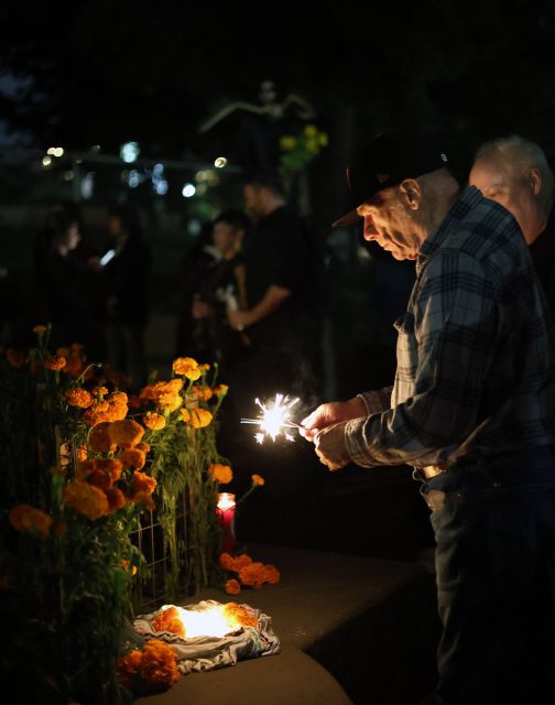 A man holds a lit sparkler near marigolds and candles at night, with others gathered nearby.