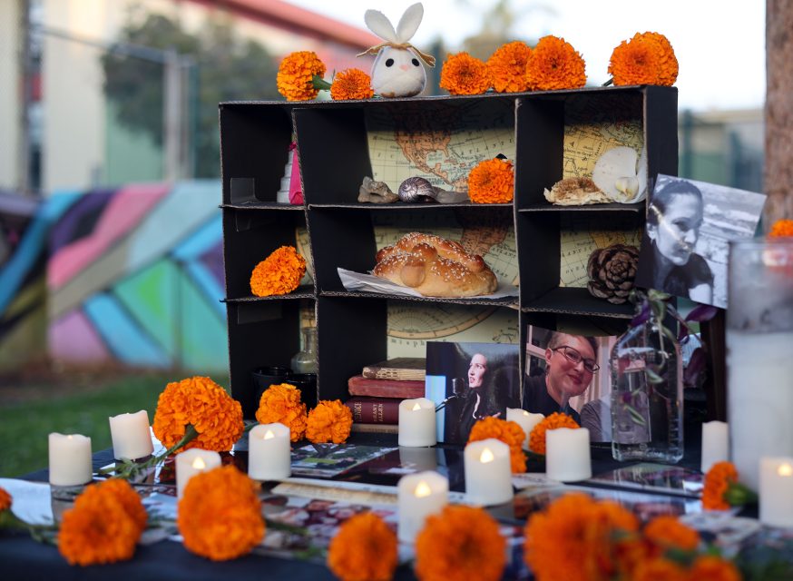 Colorful Day of the Dead altar with flowers, candles, photos, a bread roll, and a small rabbit plushie on a black shelf. Background features a mural with geometric shapes.
