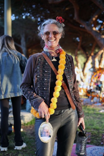 Smiling woman with sunglasses and a flower headband holds a small framed photo. She wears a decorative jacket, leather pants, and a yellow floral garland over her shoulder.
