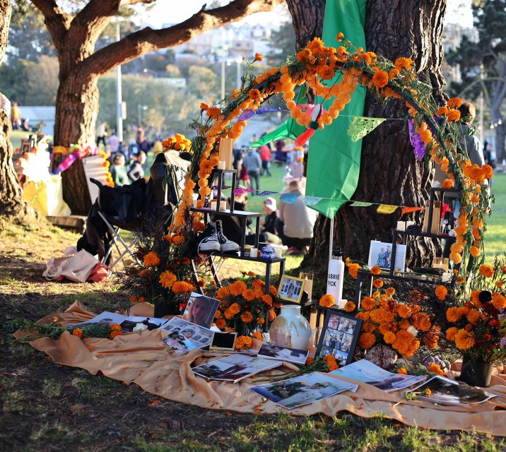 A memorial altar decorated with marigold flowers, photographs, and various items is set up outdoors under a tree during a daytime event.