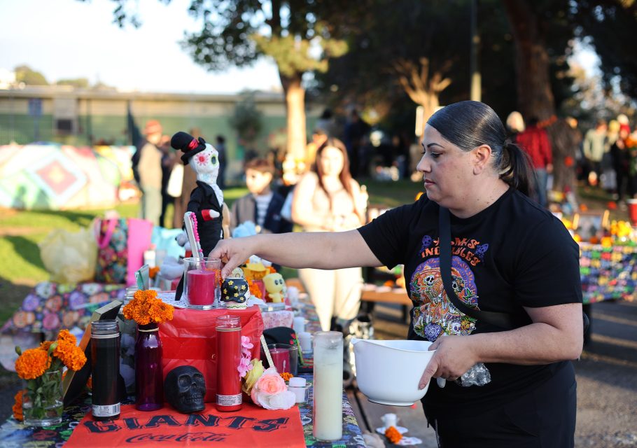 A person places items on a decorated table at a Día de los Muertos celebration, featuring candles, marigolds, and a sugar skull.