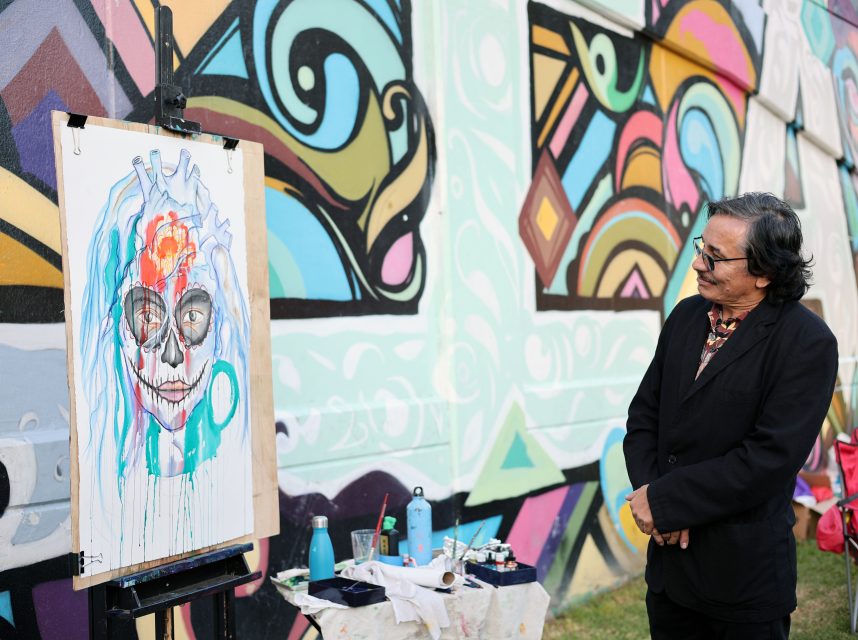 An artist in a suit stands in front of an easel with a colorful painting of a skull. He is outdoors, with a vibrant mural on the wall in the background.
