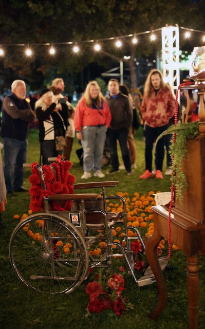 A group of people stand in front of a decorated wheelchair surrounded by marigolds, with string lights overhead in an outdoor setting.