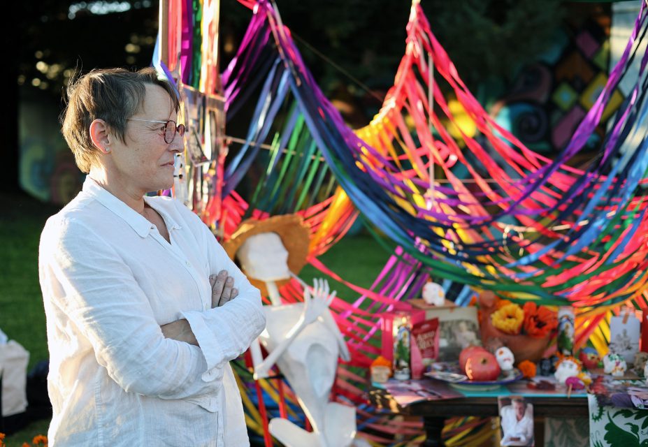 Person in a white shirt stands with arms crossed beside a vibrant art installation with ribbons and various objects.