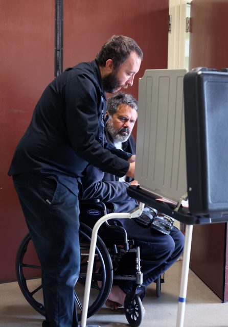A seated man in a wheelchair votes at a booth while another man stands beside him, providing assistance.