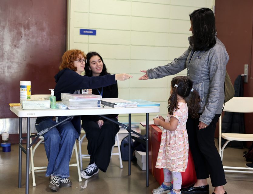 Two women sit at a table distributing items to a standing woman and a child in a community center setting.