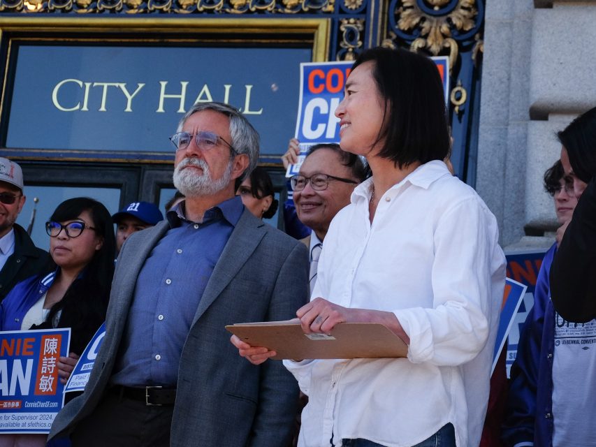 A group of people stand outside City Hall, holding signs and a clipboard, possibly for a political event or announcement.