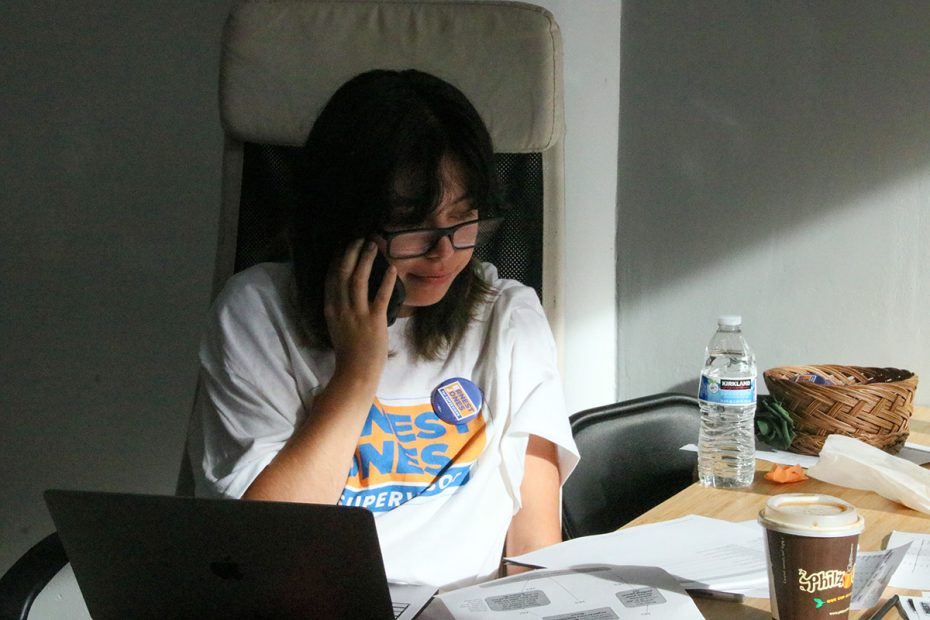 A person sits at a table, chatting on the phone amid stacks of papers, a laptop, and their trusty coffee cup. The upcoming election is clearly demanding their undivided attention.