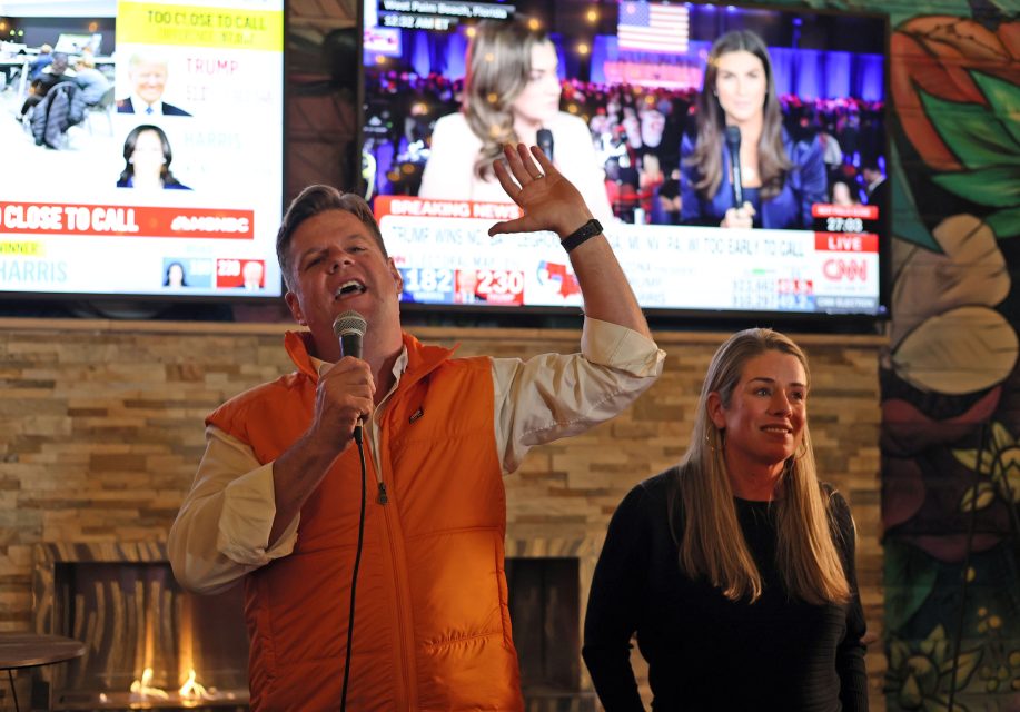 A man in an orange vest speaks into a microphone while gesturing passionately about the election, standing next to a woman. In the background, TV screens display news coverage and up-to-the-minute election updates.