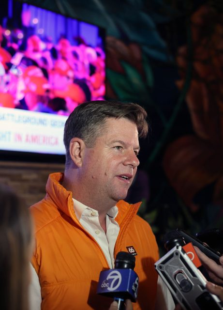 A man in an orange vest is speaking to reporters about the upcoming election. Multiple microphones are pointed at him, capturing every word. A TV screen in the background displays blurred imagery related to the event.