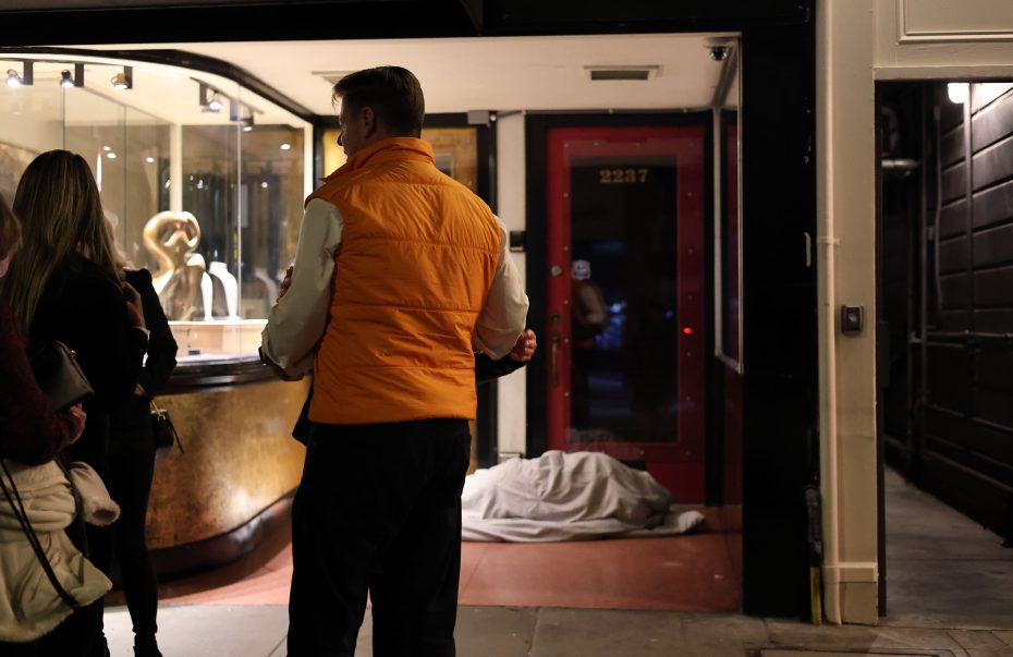 A man in an orange vest stands outside a store window, eyeing posters for the upcoming election, near a person wrapped in a blanket on the ground.