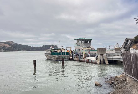 A ferry docked at a pier on a cloudy day, with hills visible in the background. Passengers are boarding or disembarking.