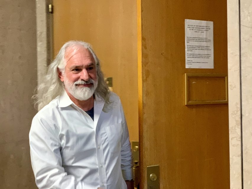 A man with long gray hair and a beard is walking through a doorway. He is wearing a white shirt and smiling.