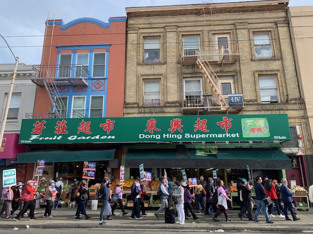 People walking on a street in front of Dong Hing Supermarket with a green sign. The building is beige with red and blue trim.