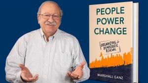 Person gesturing beside a book titled "People Power Change" by Marshall Ganz, featuring an illustration of a crowd on the cover.