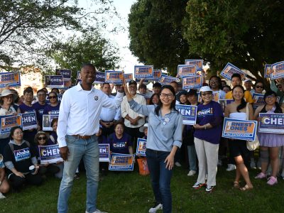 A group of people outdoors holding campaign signs for various candidates, including Ernest Jones and Chiyanne Chen. Two individuals stand in front with one raising their arm.