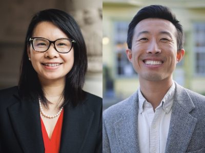 Two people side by side, both smiling. The person on the left wears a black blazer and glasses. The person on the right wears a gray jacket. Neutral background.