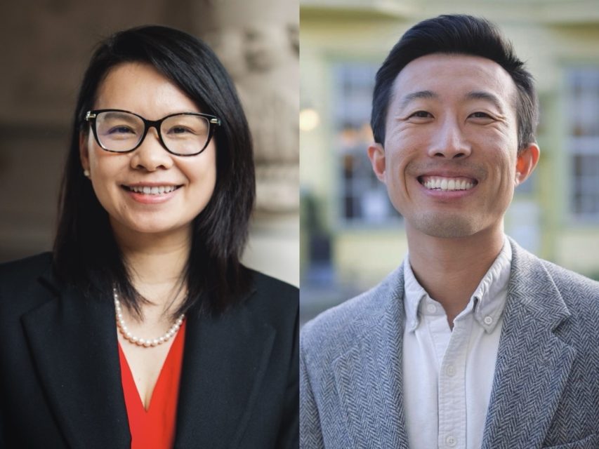 Two people side by side, both smiling. The person on the left wears a black blazer and glasses. The person on the right wears a gray jacket. Neutral background.