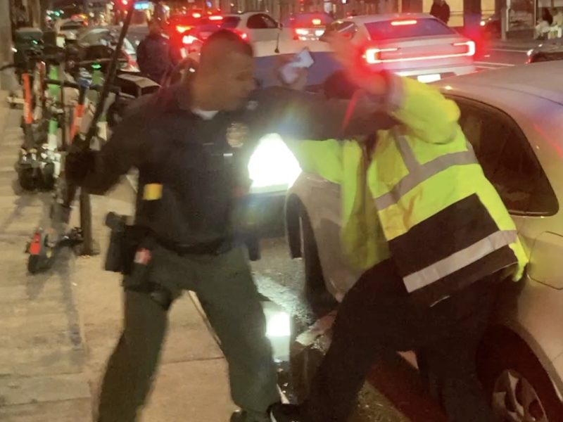 A man in a police uniform interacts with a person in a high-visibility jacket on a city street at night, with cars and scooters in the background.
