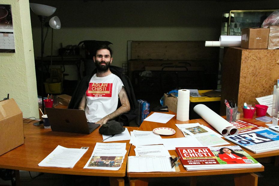 A person is seated at a cluttered wooden table, election campaign materials strewn around, wearing a shirt that reads "Adlah Chisti.