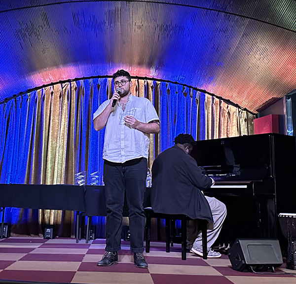 A man stands on stage holding a microphone, while another person plays the piano. The stage is lit with colorful lights and draped with curtains.