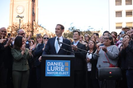A man stands at a podium labeled "Daniel Lurie Mayor-Elect," surrounded by applauding people in an outdoor setting. A clock tower is visible in the background.