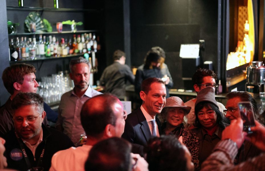 A group of people gather in a dimly lit bar, some holding drinks. A person in a suit stands centrally among them.