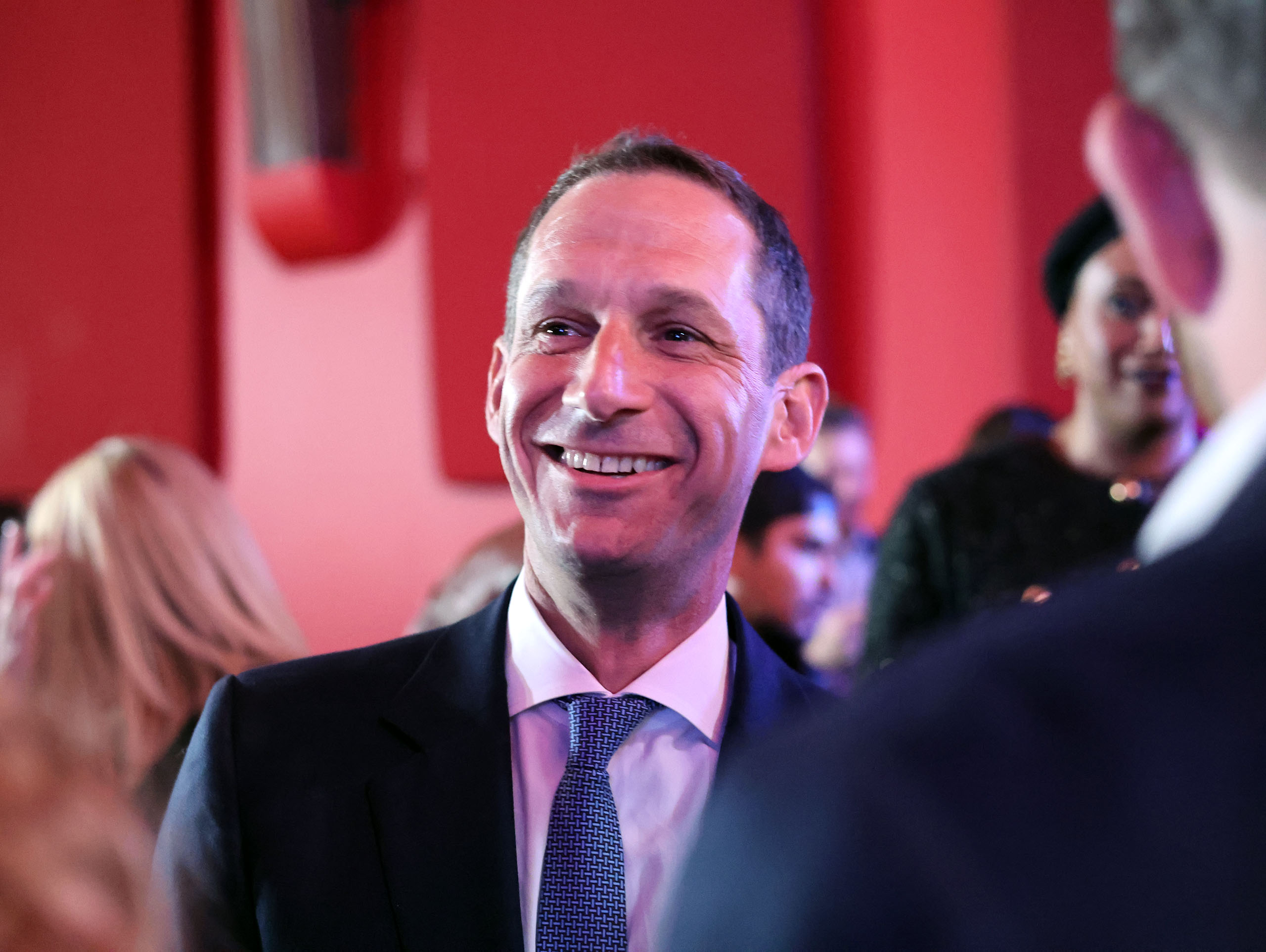 A man in a suit and tie smiles in a crowded room with a red background.