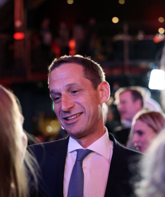 A person in a suit and tie smiles while talking to someone in a dimly lit, crowded setting.