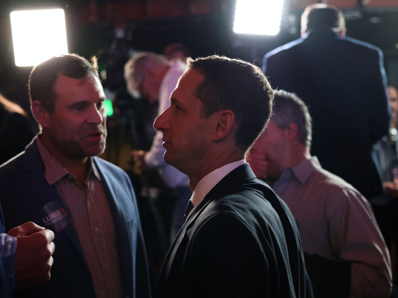A man in a suit stands in profile under bright lights, surrounded by people talking in a dimly lit space.