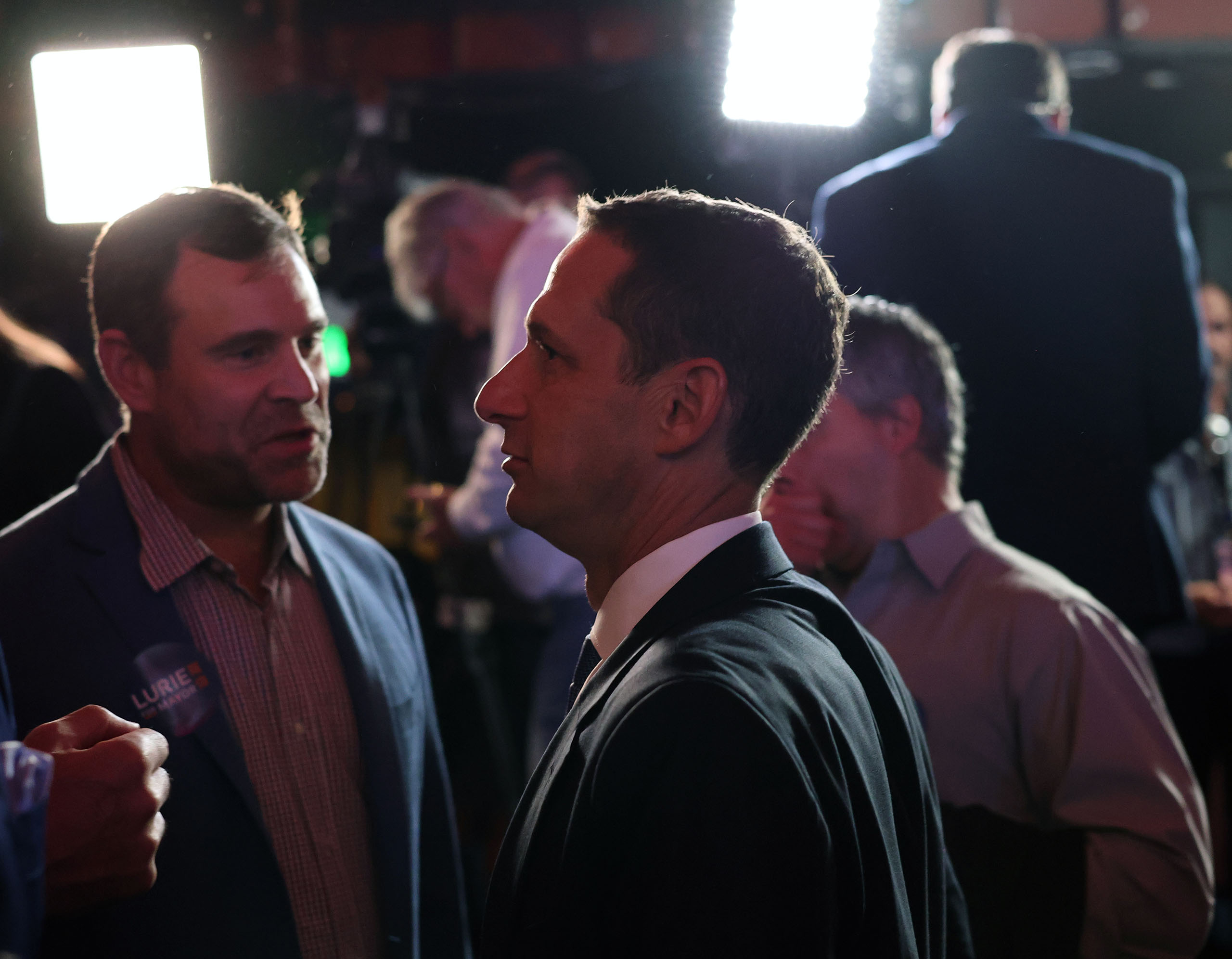 A man in a suit stands in profile under bright lights, surrounded by people talking in a dimly lit space.