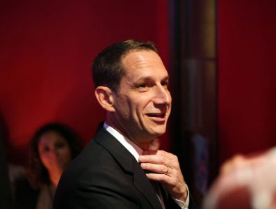 Daniel Lurie in a suit adjusts their tie, standing in front of a red wall.