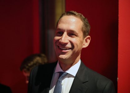 A man in a suit and tie smiles while standing against a red background.
