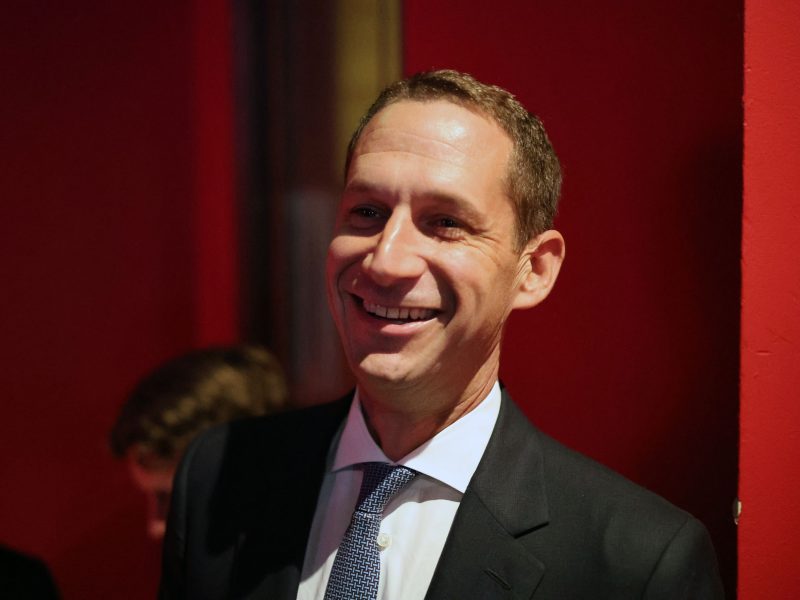 A man in a suit and tie smiles while standing against a red background.