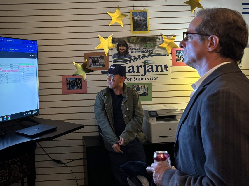 Two people in an office look at a computer screen displaying data. A can and campaign poster are visible.