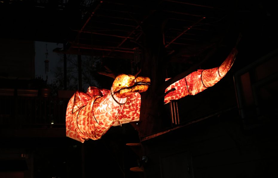 Illuminated dinosaur sculpture with a red, patterned fabric, displayed outdoors at night.