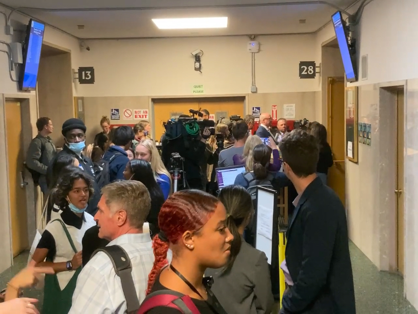 A crowded hallway with people gathered around a doorway, some with cameras and equipment.