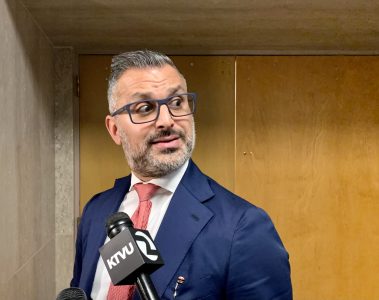 Man in a blue suit and red tie stands in front of a wooden door, holding a microphone labeled "KTVU" while looking to the side.