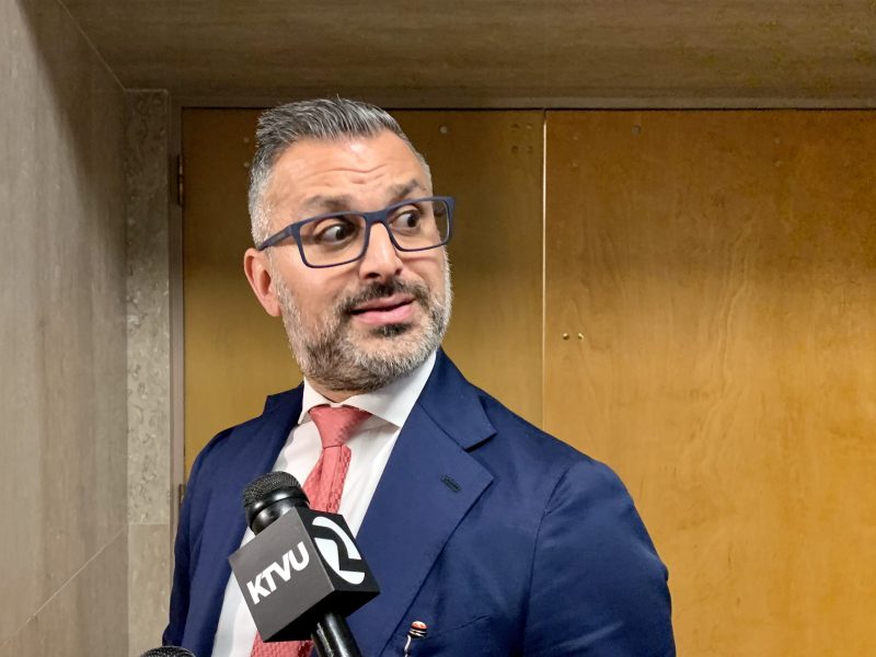 Man in a blue suit and red tie stands in front of a wooden door, holding a microphone labeled "KTVU" while looking to the side.