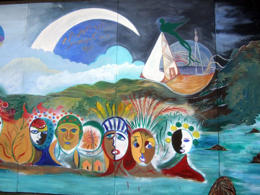 Colorful mural depicting abstract faces, a crescent moon, and a boat on a turquoise river with a cloudy sky.