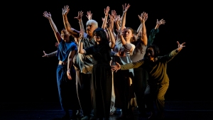 A diverse group of people stand together on stage, arms raised, with a dark background.