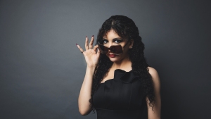 A woman in a black dress and curly hair peeks over dark sunglasses against a gray background.