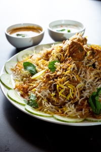 A plate of biryani garnished with cucumber slices and herbs, accompanied by two bowls of sauce on the side.