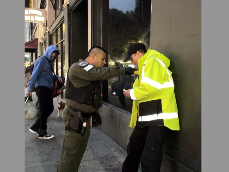 A security officer in a dark uniform checks the ID of a person in a bright yellow jacket on a city sidewalk. Another person walks by in the background.
