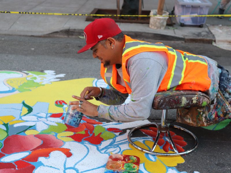A man in an orange safety vest paints a colorful mural on the street while lying on a wheeled seat.