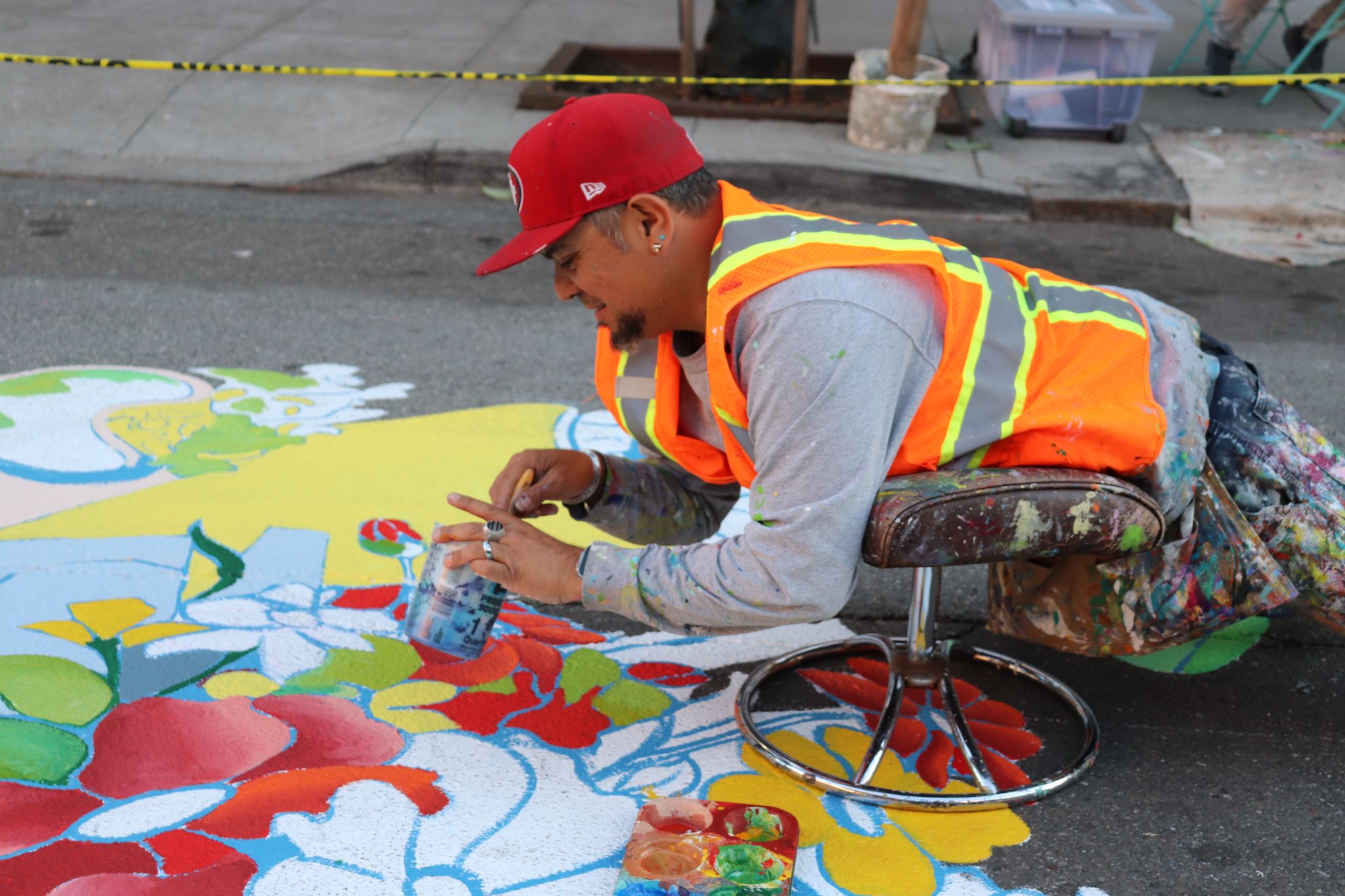 A man in an orange safety vest paints a colorful mural on the street while lying on a wheeled seat.